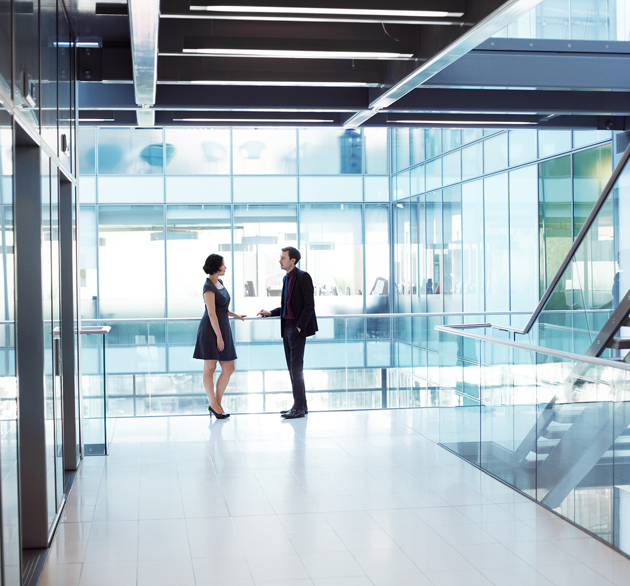 Two professionals in an office setting have a conversation while leaning on a glass railing.